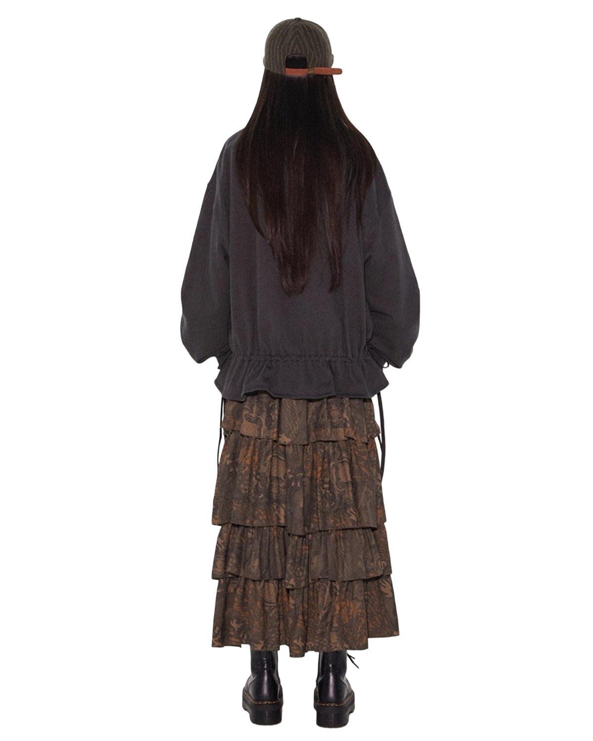 Person wearing a dark jacket and tiered brown skirt with a cap on a white background