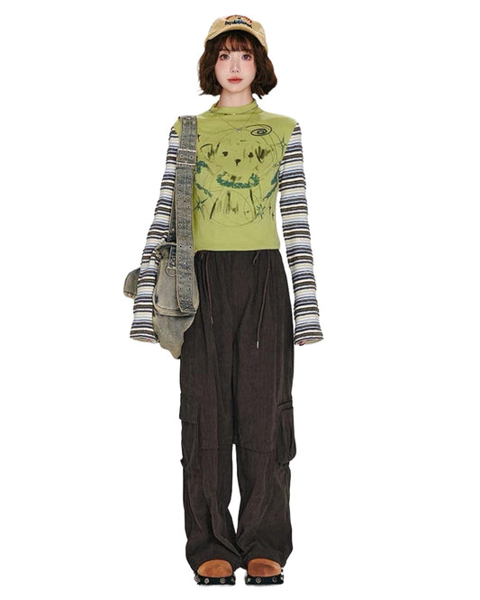 Person wearing a green graphic t-shirt, striped long-sleeve shirt, dark pants, and a cap on a white background