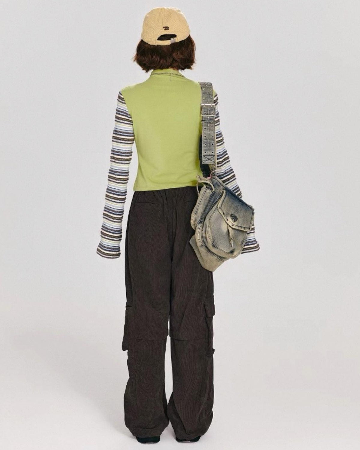 Person wearing a green vest, striped shirt, and dark pants with a backpack on a white background Back View