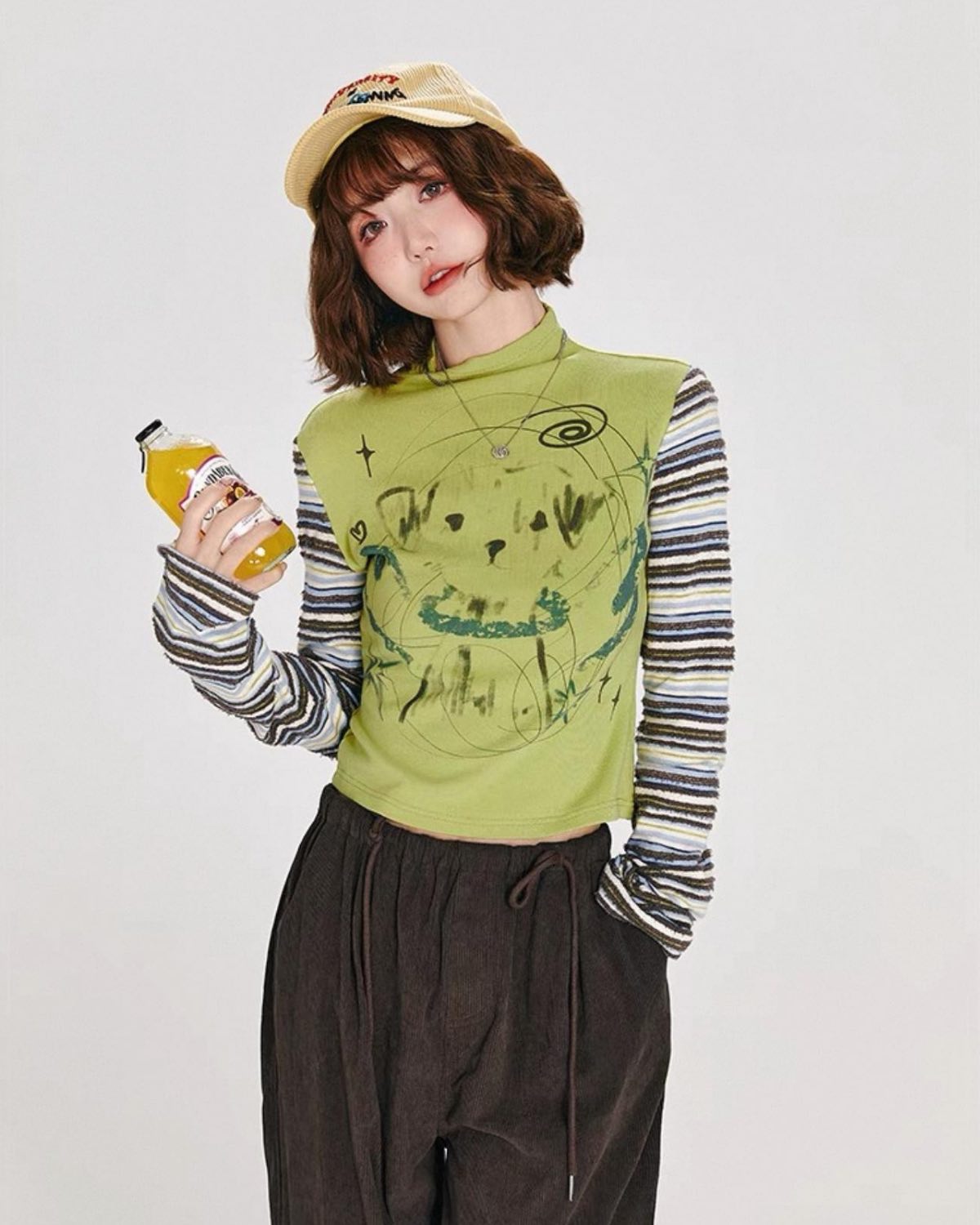 Person wearing a green graphic sweater, striped shirt, and brown pants holding a drink.