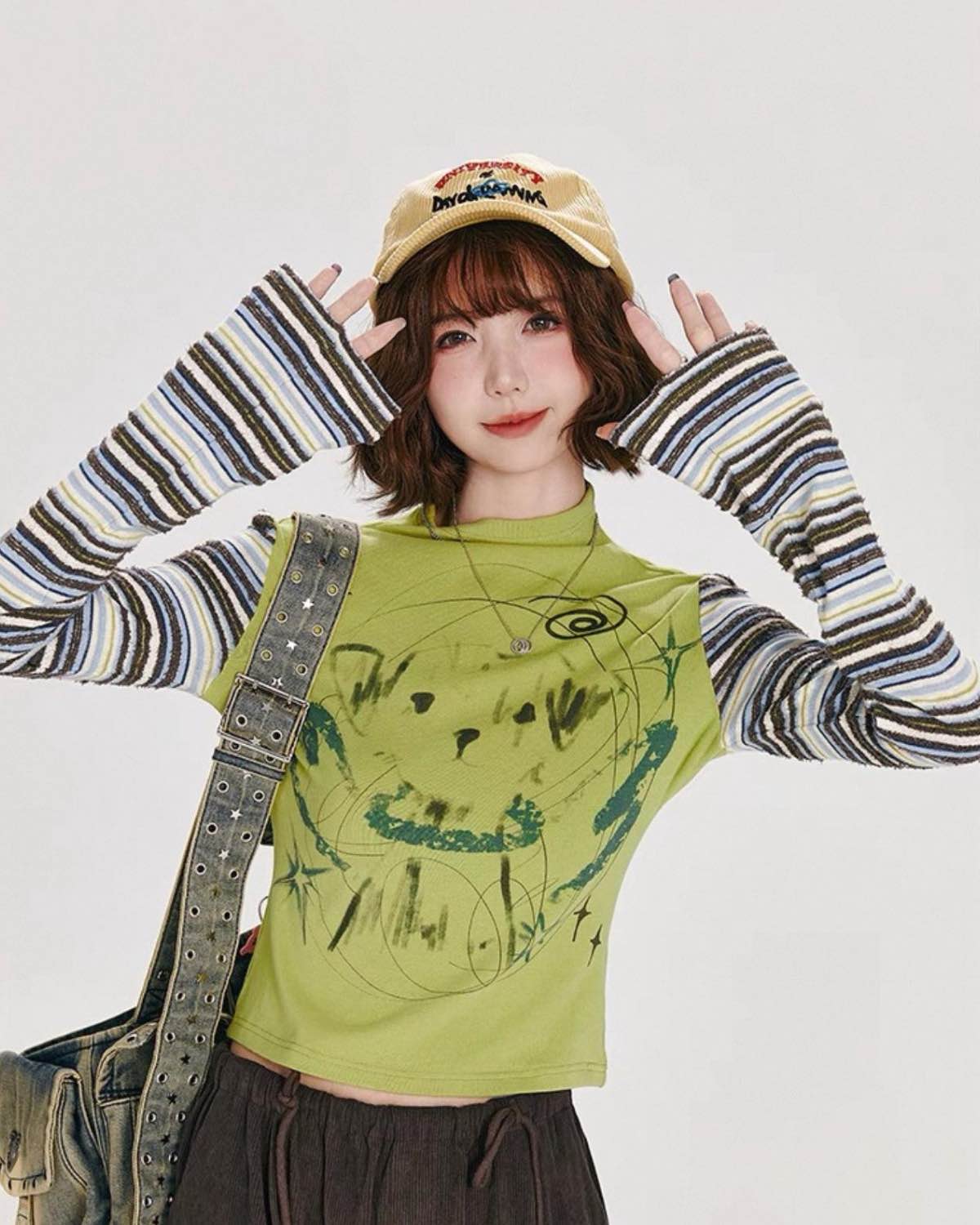 Person wearing a green graphic t-shirt with a striped long-sleeve shirt underneath, holding a beige cap.