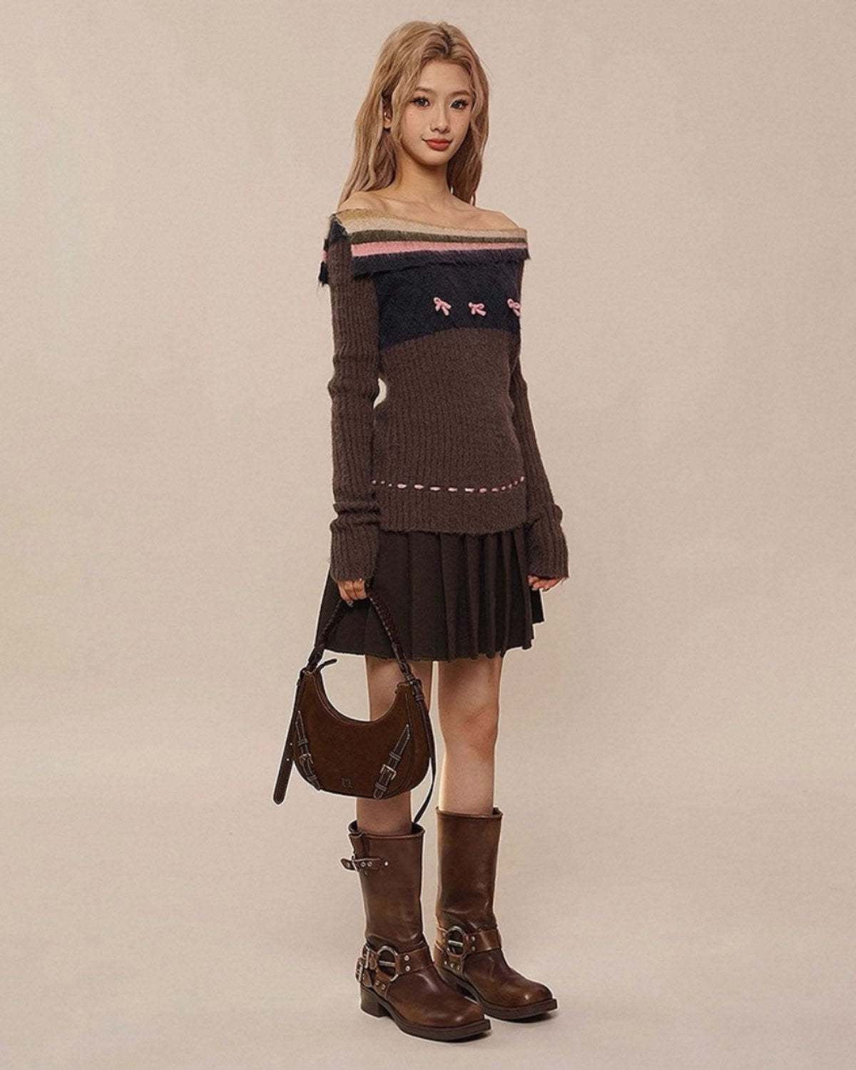 Woman wearing a brown off-shoulder sweater, black pleated skirt, and brown boots against a beige background
