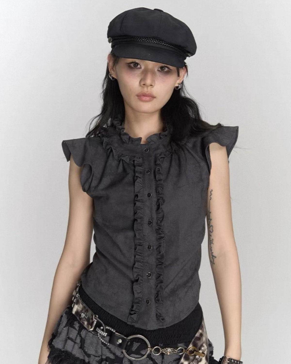 Woman wearing a dark gray sleeveless top with ruffled details and a black cap on a plain background