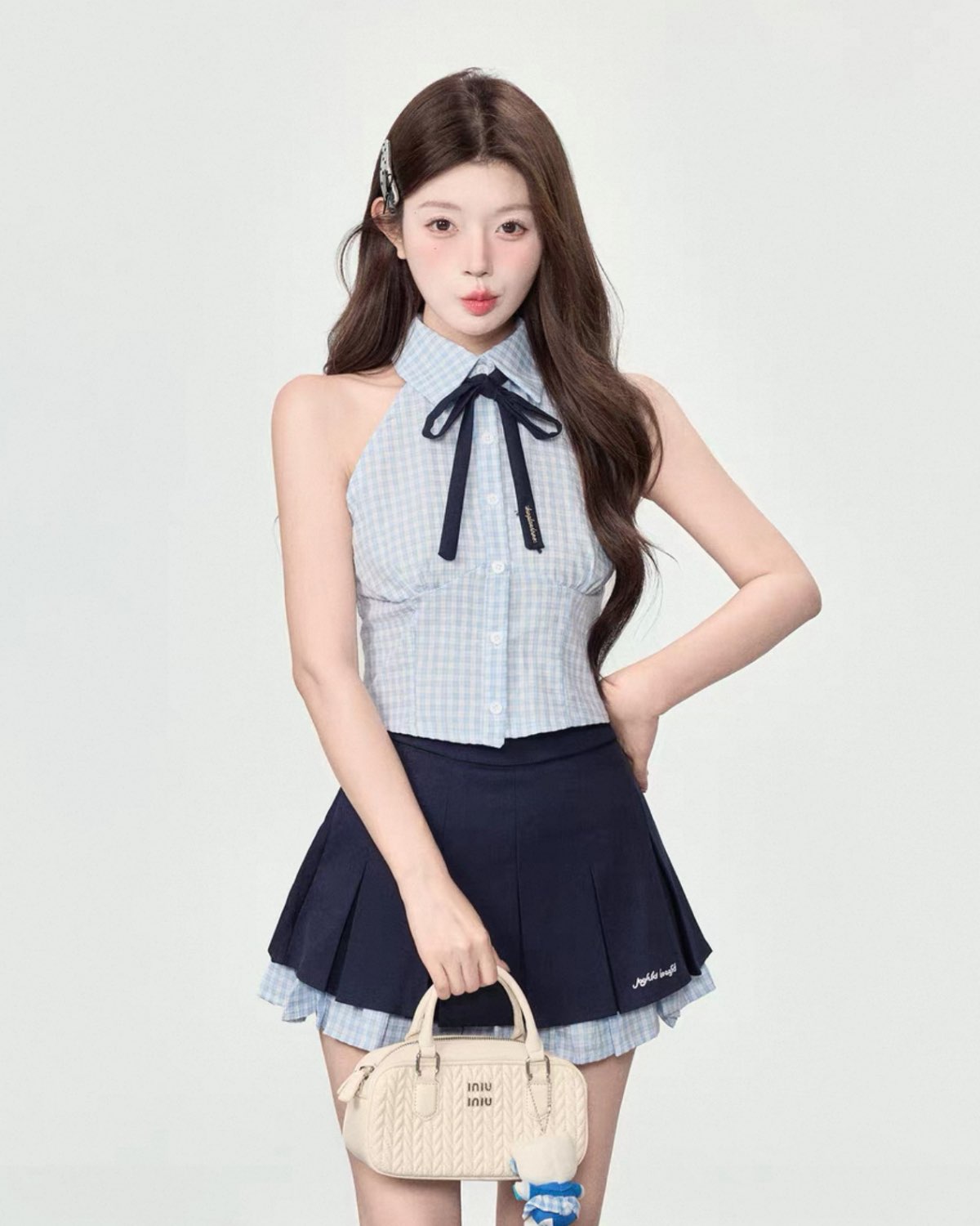 Woman wearing a checkered top with a bow and pleated skirt, holding a beige handbag on a white background