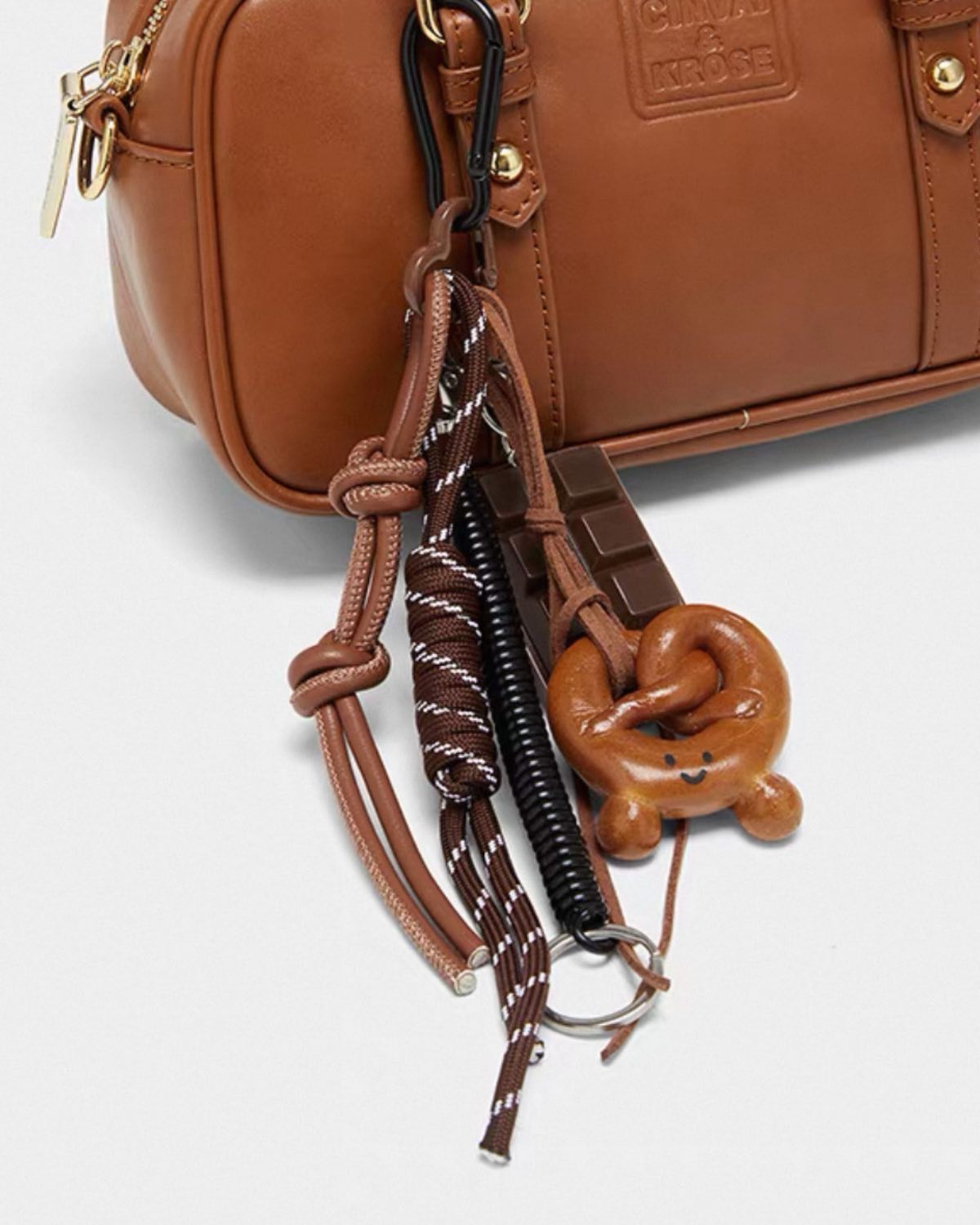 Brown leather bag with decorative keychain featuring a pretzel charm on a light gray background