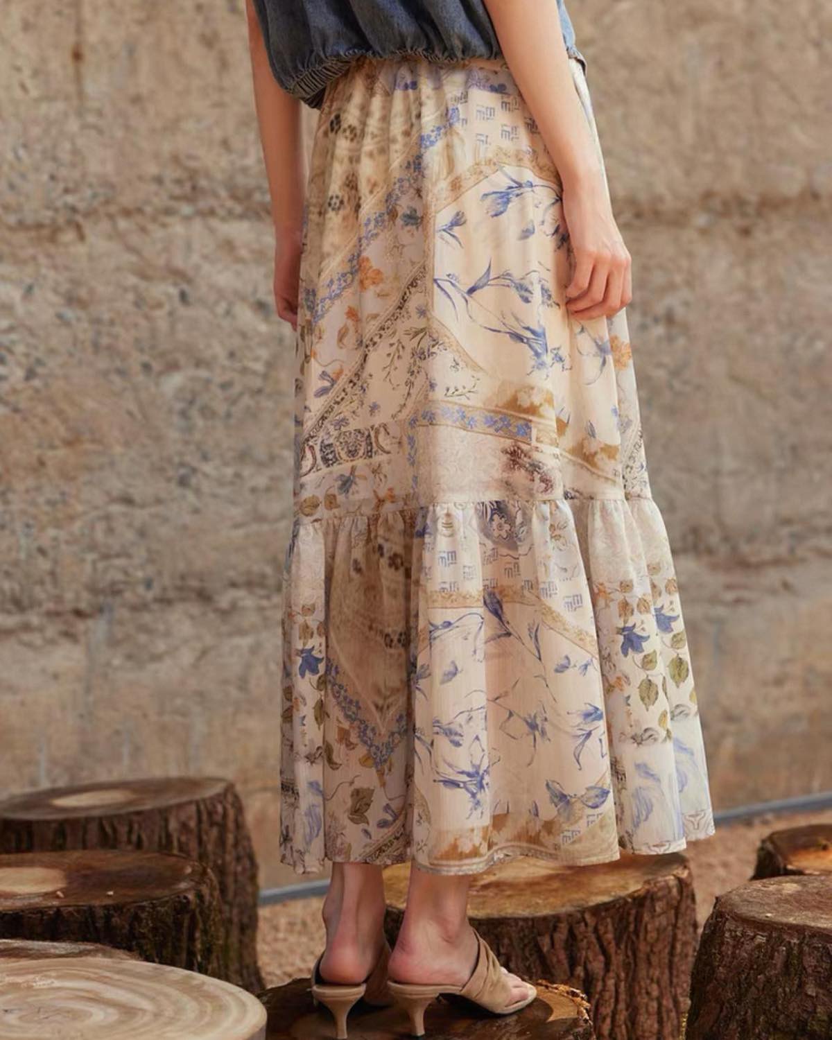 Person wearing a floral skirt standing on wooden stools against a stone wall.