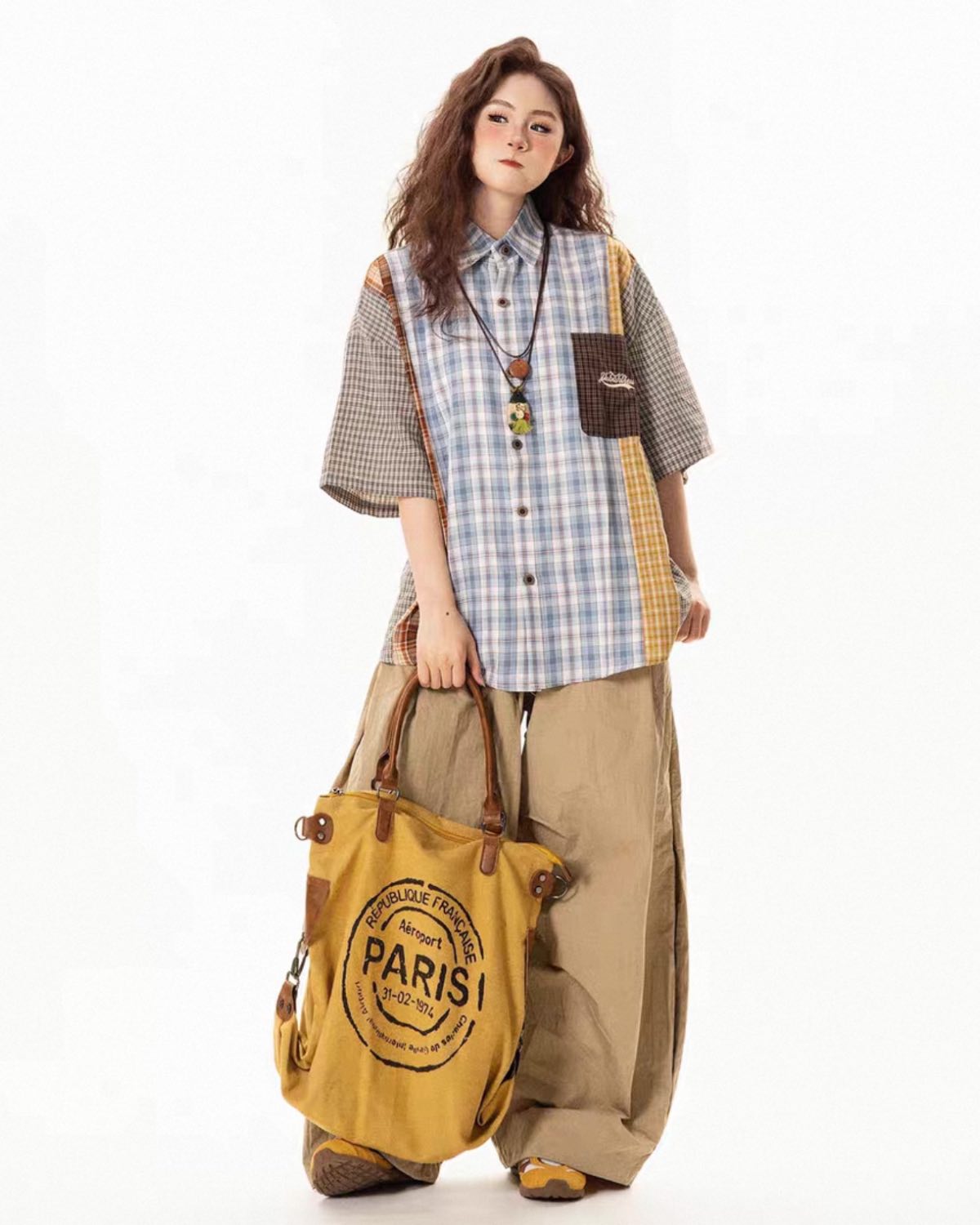 Woman holding a yellow bag with 'Paris' printed on it, wearing a plaid shirt and beige pants.
